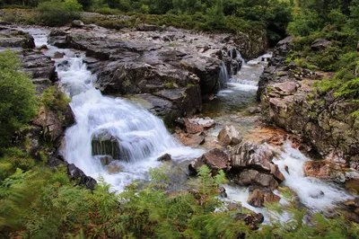 A long exposure photograph of a river cascading over dark rocks and through multiple waterfalls in a green, forested valley.