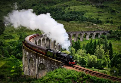 A black steam train with red carriages crosses a curved stone viaduct, emitting white smoke, set against a lush green mountainous landscape.