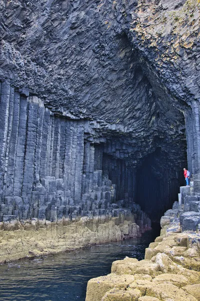 A person stands at the entrance of Fingal's Cave, a sea cave with prominent hexagonal basalt columns forming its walls and ceiling, with dark water in the foreground.