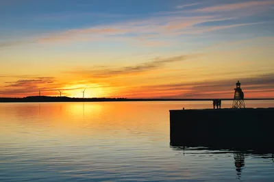 A wide shot of a sunset over water, with a lighthouse and three people silhouetted on a harbor wall. Distant wind turbines are visible on the horizon against the orange and blue sky.