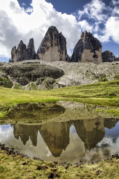 Three prominent rocky mountain peaks, known as Tre Cime di Lavaredo, rise above a green grassy landscape, with their reflections visible in a small, still lake in the foreground under a cloudy sky.