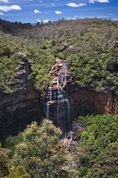 Wentworth Falls, a multi-tiered waterfall, cascades down a rocky cliff face surrounded by dense green eucalyptus forest under a bright blue sky with white clouds.