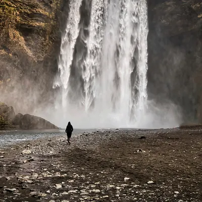 A person stands on a rocky riverbank facing a broad waterfall, with spray rising at its base.