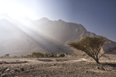 A desert mountain landscape with sunbeams, a dirt road, sparse trees, and a small village in a valley under a clear sky.