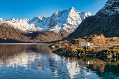 A wide shot of a Norwegian fjord with calm, reflective water. Snow-covered mountains rise dramatically in the background under a clear blue sky. A small, white house sits on a rocky, tree-dotted shore