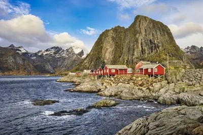 Red fishing huts on stilts along a rocky shore, with a dark blue fjord in the foreground and large, snow-capped mountains under a blue sky with clouds.