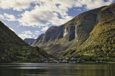 A village with colorful houses along a calm fjord, surrounded by steep, tree-covered mountains under a cloudy sky.