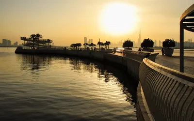 A sunset view of Dubai Creek Harbour showing a waterfront promenade with people, a spiral ramp, and the city skyline including the Burj Khalifa in the background.