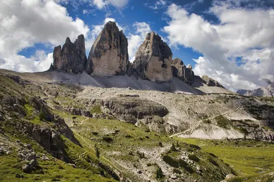 View of the Tre Cime di Lavaredo mountains in the Dolomites, Italy, with a winding hiking trail and green slopes in the foreground under a partly cloudy sky.