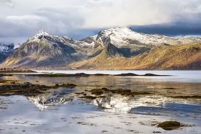 A wide landscape shot of snow-capped mountains reflecting in a calm body of water. In the foreground, dark rocks and seaweed are visible. A small village is nestled at the base of the mountains.