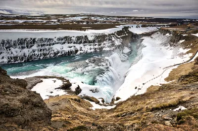 A broad waterfall plunges into a narrow rocky canyon, with snow covering the surrounding cliffs and plateau.