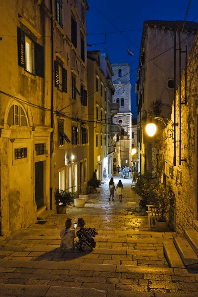 A narrow cobblestone street in an old European town at dusk, with buildings, streetlights, people, and a distant clock tower against a dark blue sky.