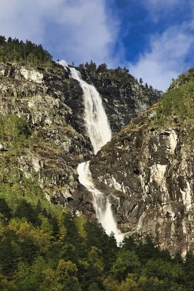 A tall, multi-tiered waterfall flows down a dark, rocky mountain covered with green pine trees. A bright blue sky with white clouds is visible above the mountain.
