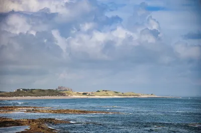 A wide view of Bamburgh Castle on a sandy beach with dunes, seen across a rocky sea foreground under a cloudy sky.