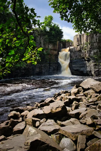 A powerful waterfall cascades into a dark river, surrounded by rocky cliffs and green trees under a bright sky.