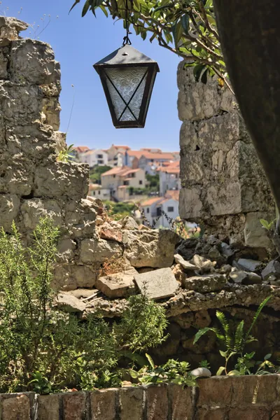 A metal lantern with patterned glass hangs from a tree branch, framed by stone ruins, with a village of white houses and red roofs visible in the background under a blue sky.