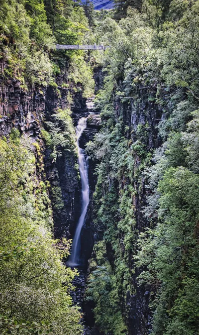 A long exposure photograph of the Falls of Measach waterfall cascading down a deep, rocky gorge covered in green trees and ferns, with a suspension bridge at the top.