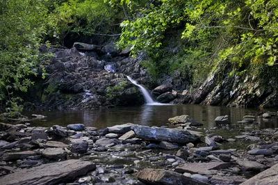 A long exposure photograph of a small waterfall cascading over dark rocks into a calm river, surrounded by green trees and moss-covered banks.