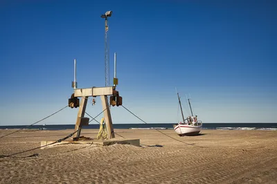 A beached white and red fishing boat sits on a wide sandy beach next to a large wooden and metal boat winch structure under a clear blue sky.