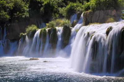Multiple waterfalls cascade over moss-covered rock formations into a river, surrounded by lush green trees and bushes on a sunny day.