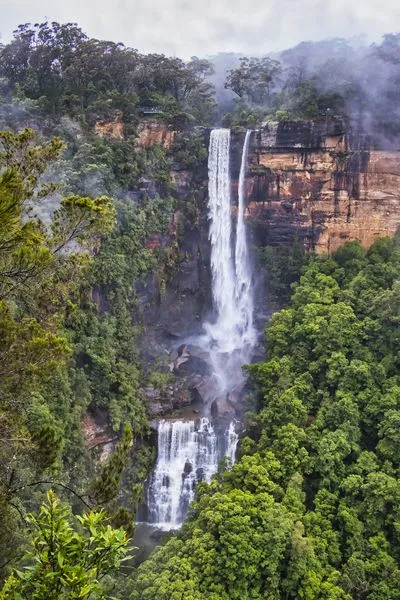 A tall waterfall cascades down a rugged cliff face into a misty pool, surrounded by dense green forest. A small viewing platform with people is visible on the upper left cliff.