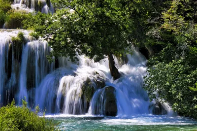 A multi-tiered waterfall cascades over rocks surrounded by dense green trees and vegetation, with clear blue water at the base.