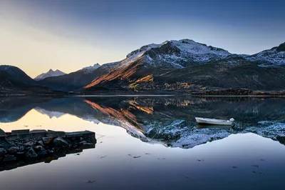 A white boat floats on a calm fjord reflecting snow-capped mountains under a clear sky. Rocks and a concrete structure are visible in the left foreground.