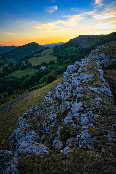 A panoramic view of a valley at sunset, featuring a rocky ridge in the foreground, green hills, trees, and a distant castle under a colorful sky.