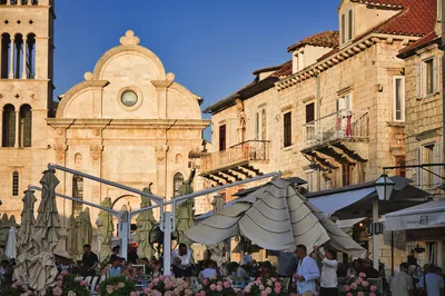 An outdoor restaurant with people dining under large umbrellas in a historic town square, with a stone church and bell tower in the background.