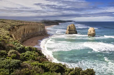 Coastal landscape with tall cliffs, a sandy beach, and two large rock stacks in the ocean with waves breaking around them.