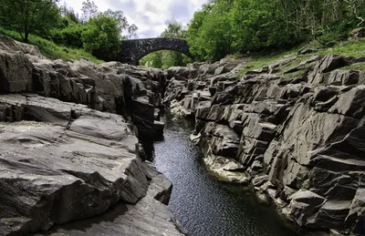 A narrow stream flows through a deep, rocky gorge with steep dark grey rock walls, leading to a stone arch bridge in the distance. Green trees line the top of the gorge.