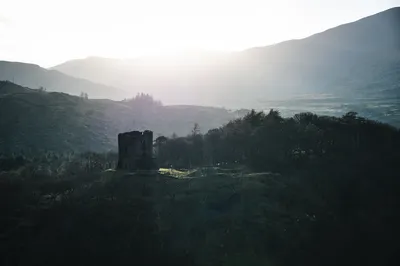 A dark, ruined castle stands on a hill, silhouetted against a bright, hazy sky with distant mountains and trees.