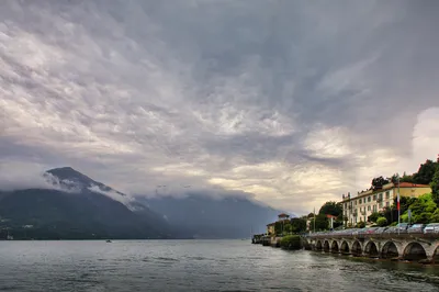 View of Lake Como with mountains partially obscured by clouds, a town with buildings and an arched promenade along the shore, and a small boat on the water.