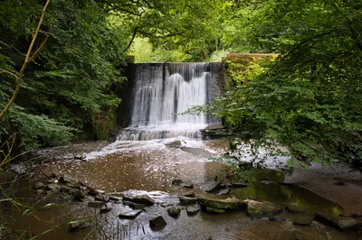 A long exposure photograph of a waterfall flowing over a man-made stone wall into a shallow river, surrounded by dense green forest foliage.