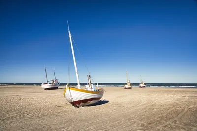 Several small, colorful fishing boats are pulled up onto a wide sandy beach, with the ocean and a clear blue sky in the background.