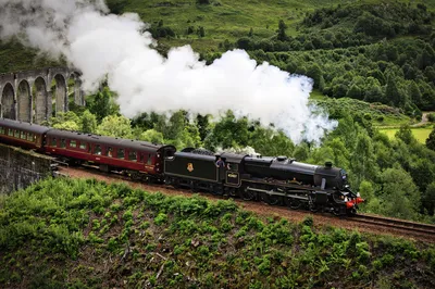 A black steam train with red carriages emits white smoke as it crosses a stone viaduct, surrounded by lush green hills and trees.
