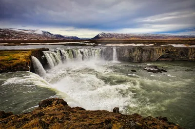 A wide waterfall spills over a rocky ledge into churning water, with snow-dusted mountains in the distance.