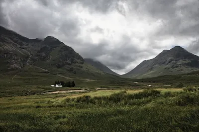 A small white house with trees in a green valley surrounded by large mountains under a cloudy sky.