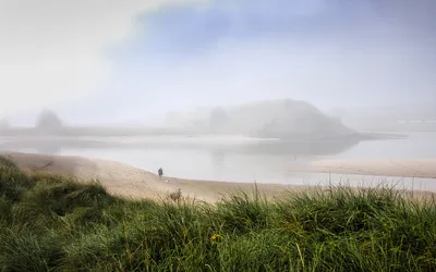 A person walks a small dog on a wide sandy beach at low tide, with green grass in the foreground and a body of water reflecting a distant, fog-shrouded island with a cross.