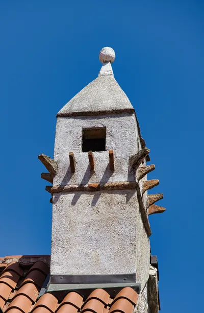 A white stucco chimney stack with a pointed roof and a spherical finial against a bright blue sky, with terracotta roof tiles visible below.