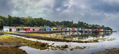 Panoramic view of a row of brightly coloured houses along a waterfront with boats, trees, and hills under a cloudy sky in Tobermory, Scotland.