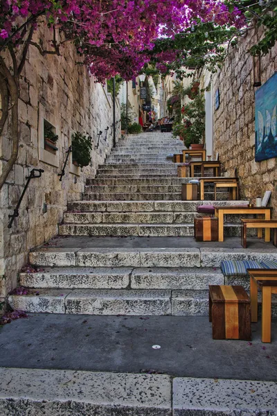 A narrow stone street with steps leading uphill, lined with cafes, tables, and chairs, and covered by a canopy of purple bougainvillea flowers.