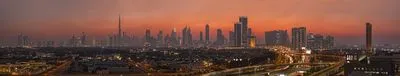 Panoramic view of Dubai's city skyline at sunset. The sky is orange and red, with numerous illuminated skyscrapers, including the Burj Khalifa. Highways in the foreground show light trails from moving