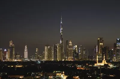 Nighttime panoramic view of the Dubai skyline, showcasing the illuminated Burj Khalifa and numerous other skyscrapers against a dark sky. A mosque is visible in the foreground.