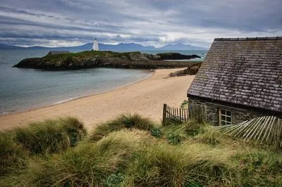 A wide shot of a sandy beach curving towards a small rocky island with a white lighthouse and a small building. In the foreground, a stone cottage with a tiled roof and wooden fence is surrounded by t