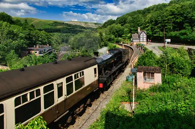 A black steam train with brown and white carriages at a station platform, surrounded by a lush green valley, river, and hills under a blue sky with white clouds. A traditional building is nearby.