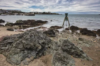 A rocky beach with a large metal tidal bell sculpture in the water. In the background, a small harbor with boats and houses lines the coast under a cloudy sky.