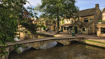 A scenic view of Bourton-on-the-Water in the Cotswolds, featuring a stone arch bridge over a calm river, traditional stone buildings, and lush green trees under a bright sky.