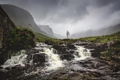 A waterfall flows over dark rocks in a rugged landscape with a single tree, misty mountains, and a stone wall under a cloudy sky.