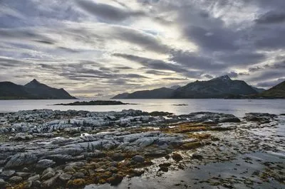 A wide landscape shot of a rocky coastline with calm water, distant mountains, and a dramatic cloudy sky in Lofoten, Norway. The foreground features light-colored, layered rocks and some seaweed.